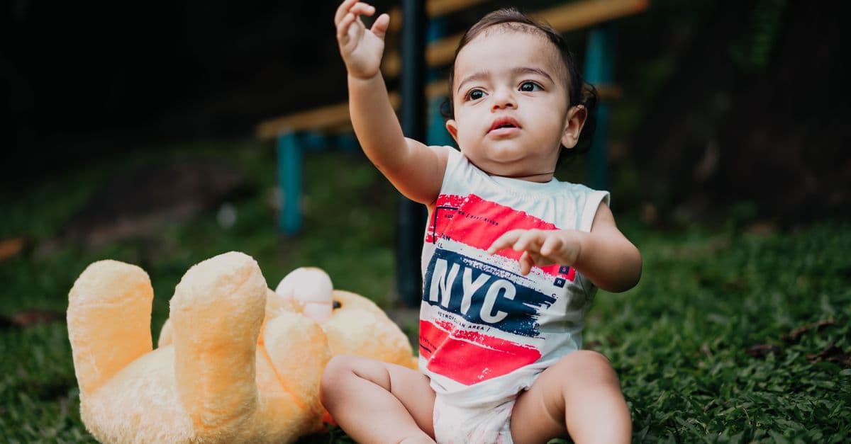 Cute baby with a teddy bear sitting on grass, wearing 'NYC' tank top.
