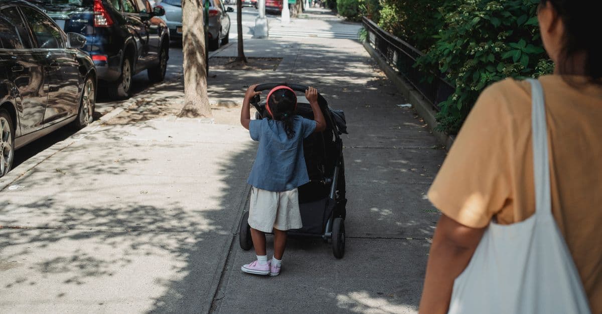 Back view of faceless crop ethnic mother with bag walking on sunny pathway with anonymous girl rolling baby stroller during promenade